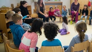 Kinder und Erzieherinnen sitzen in einer Kita im Kreis. In Rheinland-Pfalz haben immer mehr Kita-Beschäftigte keine pädagogische Ausbildung.