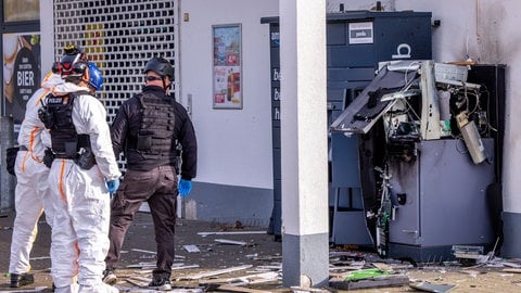 Zwei Polizisten in weißen Schutzanzügen und ein Polizist in schwarzer Sicherheitsausrüstung stehen vor einem gesprengten Geldautomaten.