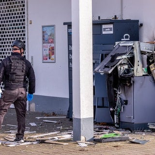 Zwei Polizisten in weißen Schutzanzügen und ein Polizist in schwarzer Sicherheitsausrüstung stehen vor einem gesprengten Geldautomaten.