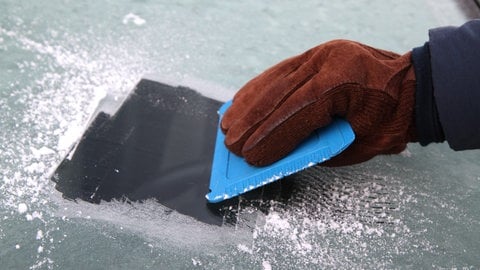 Eine Hand kratzt mit einem Eiskratzer die von Eis und Schnee festgefrorene Autoscheibe, genauerm die Frontscheibe, frei. 