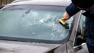 Ein Mann kratzt das Eis von der Fensterscheibe eines Autos - in höheren Lagen in Rheinland-Pfalz kann es heute Morgen zu Glatteis kommen.