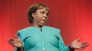 Marina Tamássy steht als Angela Merkel auf der Bühne. Sie trägt eine grüne Jacke. 