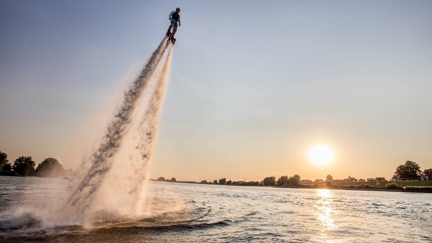 Mensch schießt vor einem Sonnenuntergang mit einem Flyboard aus dem Wasser