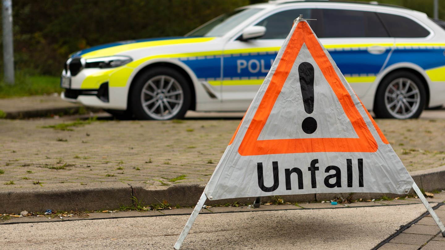 An einer Straße steht ein faltbares Warndreieck der Polizei mit der Aufschrift Unfall. Im Hintergrund steht ein Streifenwagen der Polizei.