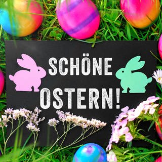 Ostergruß: Schöne Ostern! Auf einem Schild in einer Wiese mit bunten Ostereiern.