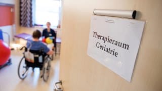 Eine Zimmertür in einer Klinik steht offen. An der Tür hängt ein Schild mit der Aufschrift "Therapieraum Geriatrie".