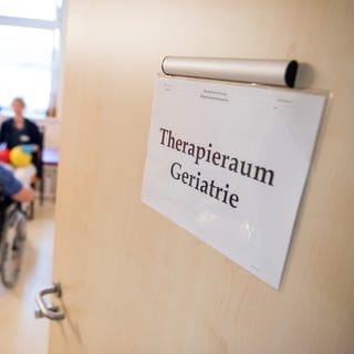 Eine Zimmertür in einer Klinik steht offen. An der Tür hängt ein Schild mit der Aufschrift "Therapieraum Geriatrie".