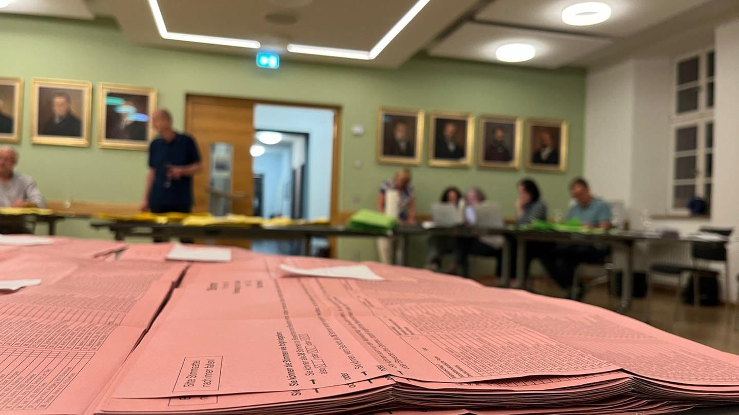 Stimmzettel liegen aufgestapelt im Vordergrund, dahinter sind Wahlhelfer in Kusel an Tischen mit dem Auszählen der Stimmen beschäftigt. (SWR)