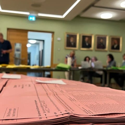 Stimmzettel liegen aufgestapelt im Vordergrund, dahinter sind Wahlhelfer in Kusel an Tischen mit dem Auszählen der Stimmen beschäftigt. (SWR)