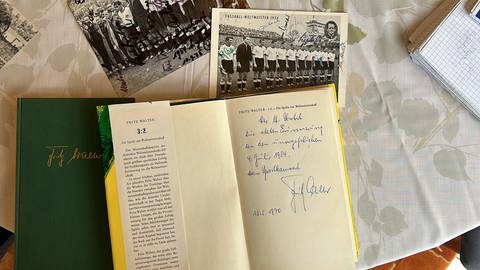 Ein aufgeschlagenes Buch mit Widmung von Fritz Walter auf der ersten Seite liegt auf dem Tisch. Darüber alte Autogrammkarten der deutschen Fußballmannschaft. (SWR)