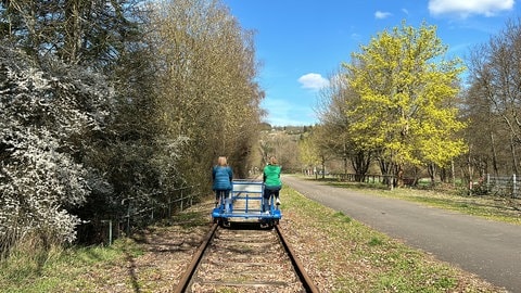 Zwei Frauen fahren auf einer Draisine durch die frühlingshafte Landschaft bei Altenglan.