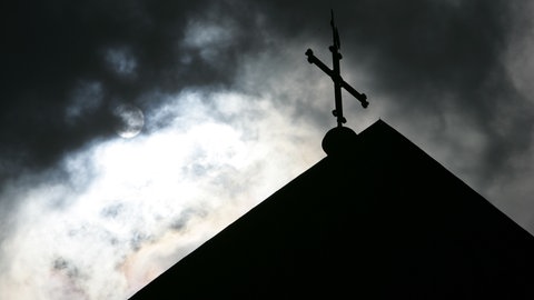 Im Gegenlicht und vor wolkenverhangenem Himmel ist die Kirchturmspitze eines Doms mit Kreuz zu sehen.