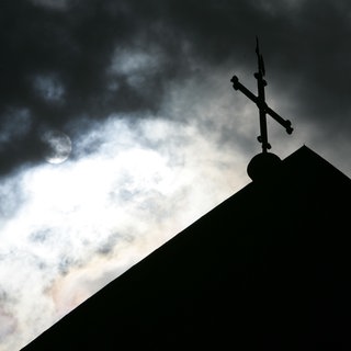 Im Gegenlicht und vor wolkenverhangenem Himmel ist die Kirchturmspitze eines Doms mit Kreuz zu sehen.