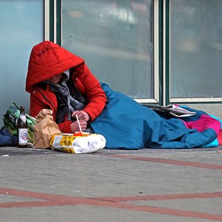 Ein Mensch liegt auf dem Boden, eingepackt in eine dicke Jacke und einen Schlafsack.In Koblenz und der Region bieten viele Vereine und Organisationen Wohnungslosen auch in diesem Winter warmes Essen und Kleidung, Übernachtungsplätze und Schlafsäcke an. 