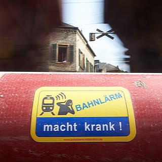 Ein Güterzug fährt am Mittelrhein unmittelbar an Wohnhäusern und an einer Schranke mit der Aufschrift "Bahnlärm macht krank" vorbei. 