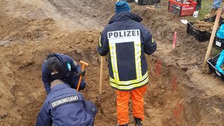 Drei Menschen in Arbeitskleidung stehen und hocken mit Schaufeln in einer Grube. Zwei von ihnen tragen Jacken mit der Beschriftung "Ordnungsamt". In der Grube auf einer Baustelle im Engerser Feld in Neuwied wurden die Leichen von vier deutschen Soldaten aus dem Zweiten Weltkrieg gefunden.