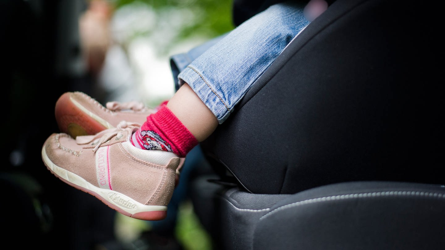 Ein Kleinkind mit braunen Schuhen und roten Socken sitzt im Autositz. Man sieht den Fußbereich und den unteren Teil des Sitzes.