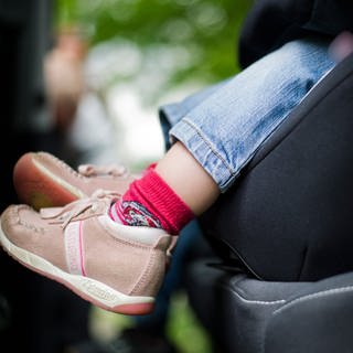 Ein Kleinkind mit braunen Schuhen und roten Socken sitzt im Autositz. Man sieht den Fußbereich und den unteren Teil des Sitzes.