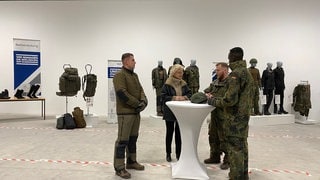 Bundesverteidigungsministerin Lambrecht steht im Bekleidungszentrum der Bundeswehr in Simmern zusammen mit Soldaten an einem Tisch. 