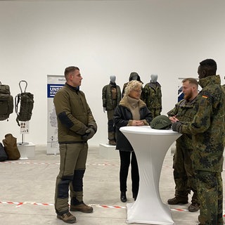 Bundesverteidigungsministerin Lambrecht steht im Bekleidungszentrum der Bundeswehr in Simmern zusammen mit Soldaten an einem Tisch. 