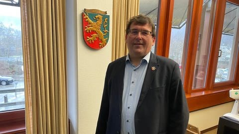 Jörg Denninghoff, Landrat des Rhein-Lahn-Kreises, steht vor einem Wappen in seinem Büro