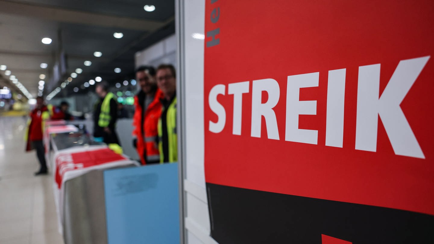 Am Flughafen Köln/Bonn steht ein Streik-Schild von Verdi und im Hintergrund sind Mitarbeiter zu sehen