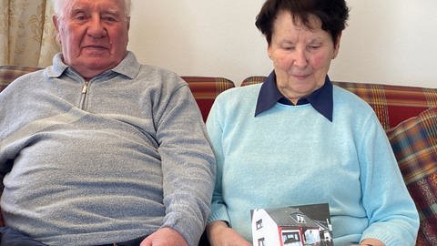 ein älteres Ehepaar sitzt beeinander, sie hält ein Foto von ihrem Haus in der Hand aus der Zeit vor der Flutkatastrophe.