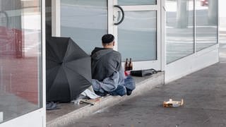 Obdachloser von hinten, sitzt vor dem Eingang eines Geschäfts