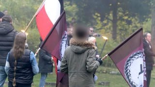 Drei Menschen halten eine Fahne bei einer rechtsextremen Veranstaltung in der Hand, darunter ein Junge in einer oliven Jacke.