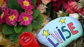 Blumen, Kerzen und ein bunt bemalter und mit "Luise" beschriebener Stein liegen am Tatort: Die Eltern der getöteten Zwölfjährigen verklagen die beiden mutmaßlichen Täterinnen vor dem Landgericht Koblenz auf Schmerzensgeld