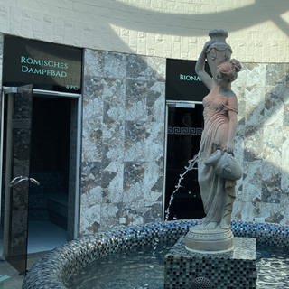 Ein Brunnen mit einer weiblichen Statue, die Amphoren trägt. Dahinter sieht man die Türen der verscheidenen Saunen und der Umkleiden.