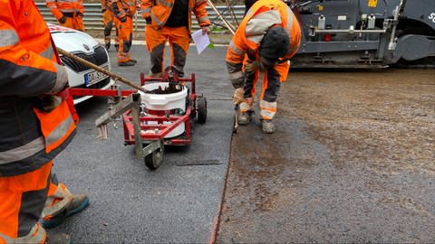 Ein Bauarbeiter pinselt die Kante zwischen dem fertigen und dem unfertigen Teil der Fahrbahn mit Klebemittel ein, damit im Anschluss asphaltiert werden kann.