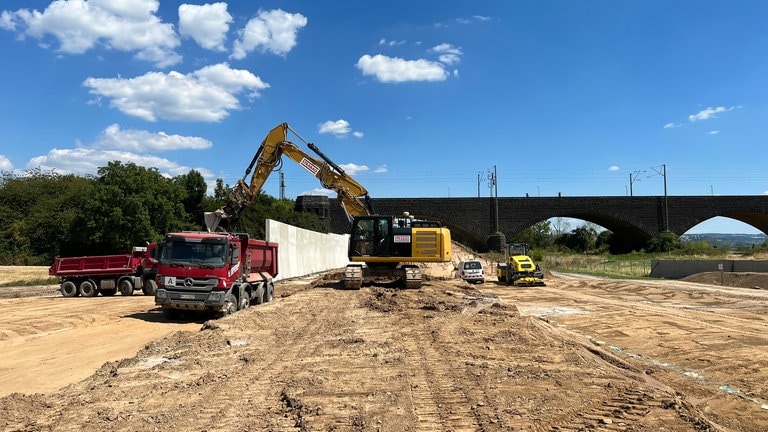 Der Rheindeich in Neuwied-Engers bei der Eisenbahnbrücke Engers-Urmitz im Wiederaufbau. Ein Bagger schaufelt Erde aus einem LKW für den Bau des Deiches. Zudem sieht man eine Betonsicherheitsmauer.