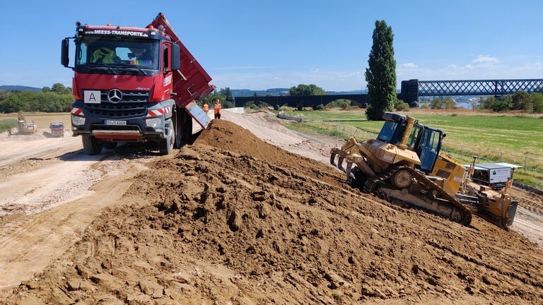 Der Rheindeich in Neuwied-Engers bei der Eisenbahnbrücke Engers-Urmitz im Wiederaufbau. Ein LKW lädt Erde ab und Baumaschinen richten das Gefälle des Deiches.