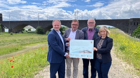 Jan Einig (Oberbürgermeister Stadt Neuwied), Wolfgang Treis (Präsident SGD Nord RLP) und Katrin Eder (Klimaschutzministerin RLP) halten die Infotafel für den sanierten Deich hoch.