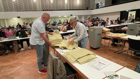 Zwei Männer stehen am Tag nach der Kommunalwahl 2024 in der Rhein-Mosel-Halle an einem Tisch mit hunderten Wahlunterlagen