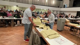 Zwei Männer stehen am Tag nach der Kommunalwahl 2024 in der Rhein-Mosel-Halle an einem Tisch mit hunderten Wahlunterlagen.