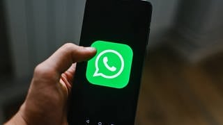 Eine männliche Hand hält ein Handy mit dem grünen Logo von WhatsApp auf dem Display