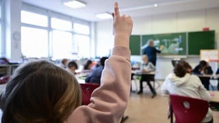 Situation im Klassenzimmer einer Grundschule, Lehrer an der Tafel, ein Kind hebt den Finger