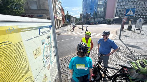 Gruppe von Radfahrern steht vor einem Schild in Koblenz, das den Rheinradweg zeigt
