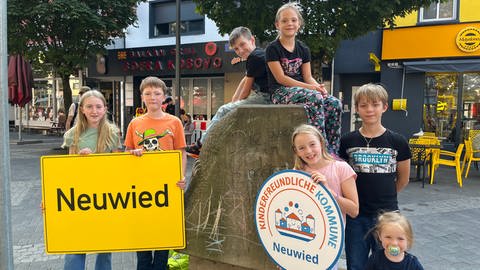 Das Kinderprinzenpaar Oberbieber und sein Gefolge stehen bei dem Kinderrechte-Denkmal in Neuwied. Sie halten ein Ortsschild und eine Plakette mit der Aufschrift "Kinderfreundliche Kommune Neuwied". Zusammen setzen sie sich für die Kinderrechte ein.