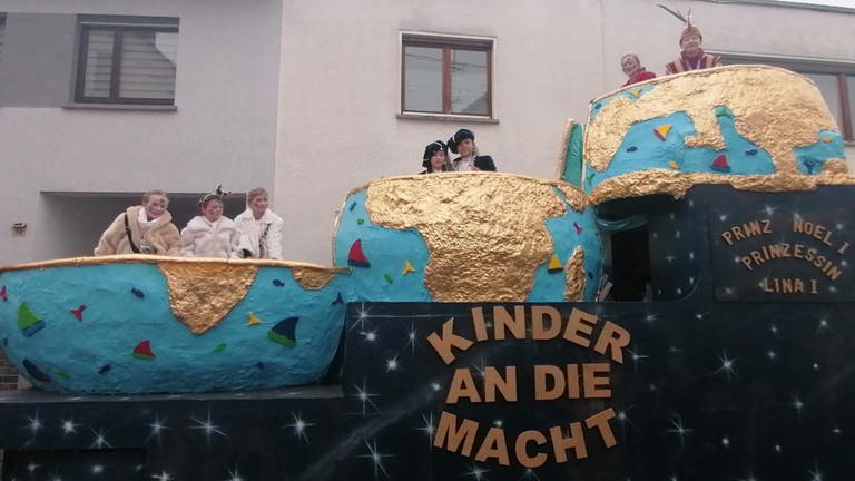 Das Kinderprinzenpaar Oberbieber und sein Gefolge fahren bei einem Karnevalsumzug auf ihrem Festwagen mit. Darauf steht: Kinder an die Macht.