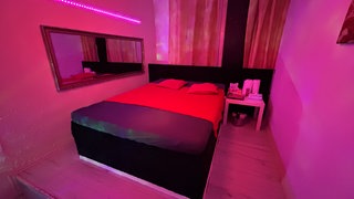 Zimmer in einer Prostituiertenwohnung in Lahnstein.Er ist spärlich beleuchtet, mit rotem Bett und Hygieneartikel auf dem Seitentisch. 