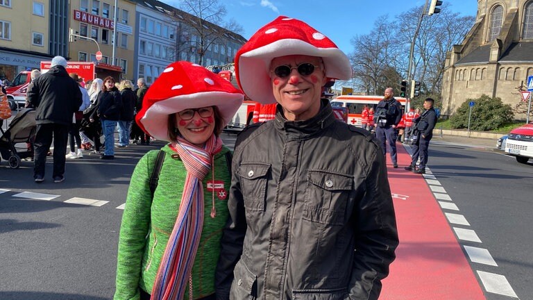 Ein Mann und eine Frau lachen mit rot-weißen Pilzkopf-Hüten beim Rosenmontagszug 2023 in Koblenz in die Kamera.