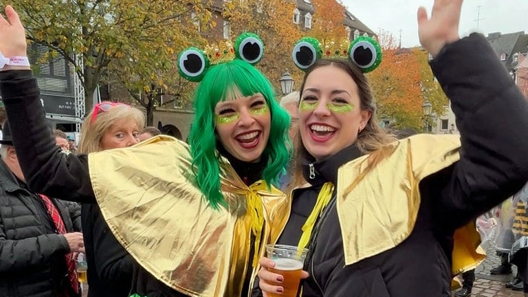 Zwei junge Frauen mit grünen Haaren und Frosch-Augen auf dem Kopf sind beim Karnevalsauftakt 2024 in Koblenz dabei. 