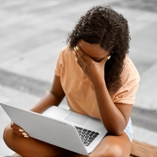 Eine junge Frau sitzt mit einem Laptop auf dem Schoss auf dem Boden. Ein Hand schlägt sie verzweifelt vor die Augen.