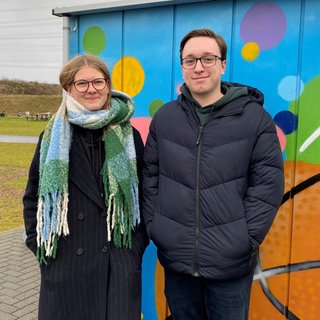 Eine junge Frau und ein junger Mann stehen vor einem bunt bemalten Container auf dem Schulgelände des Are-Gymnasiums in der Grafschaft. 