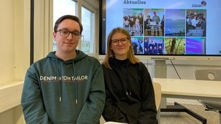 Eine junge Frau und ein junger Mann lächeln in die Kamera. Sie sitzen vor einer großen Leinwand.