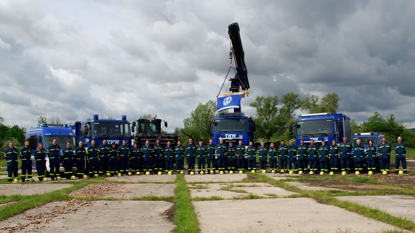 THW-Mitglieder stehen in dunkelblauen  Uniformen vor mehreren marineblauen Einsatzfahrzeugen, darunter einem Kran