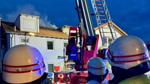 Einsatzkräfte stehen im Vordergrund. Im Hintergrund sieht man das brennende Haus.